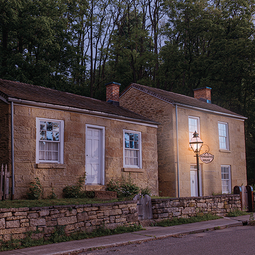 Pendarvis house at dusk, orange colored brick and white trim windows and doors. A stone wall lines the house next to the road. The lamp is lit.