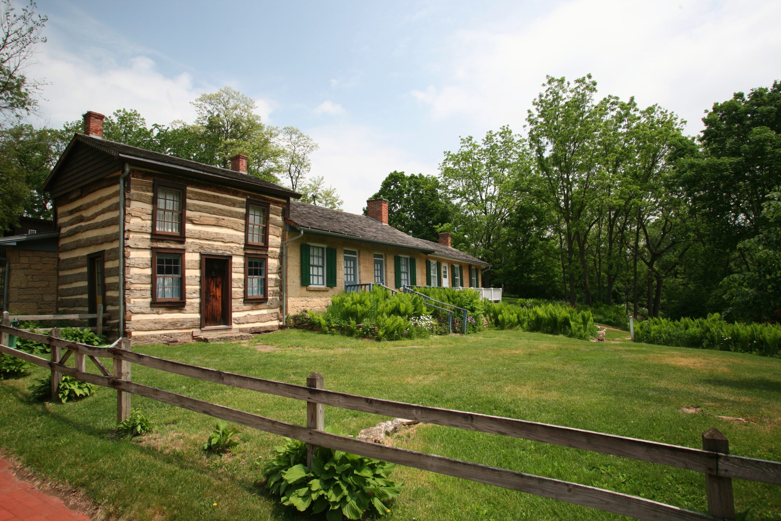 Well preserved, historic log cabin with a long structure and green shutters, surrounded by wooden fence and lush greenery. Martin Cabin, Pendarvis.