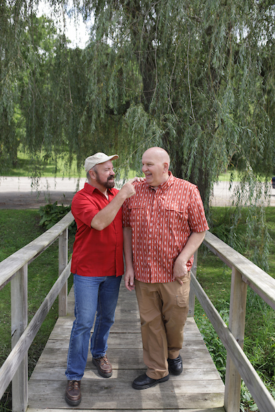 two men on a wooden bridge, one in a red shirt and cap speaking and gesturing to the other in a patterned red shirt, in what appears to be a friendly and engaging conversation.